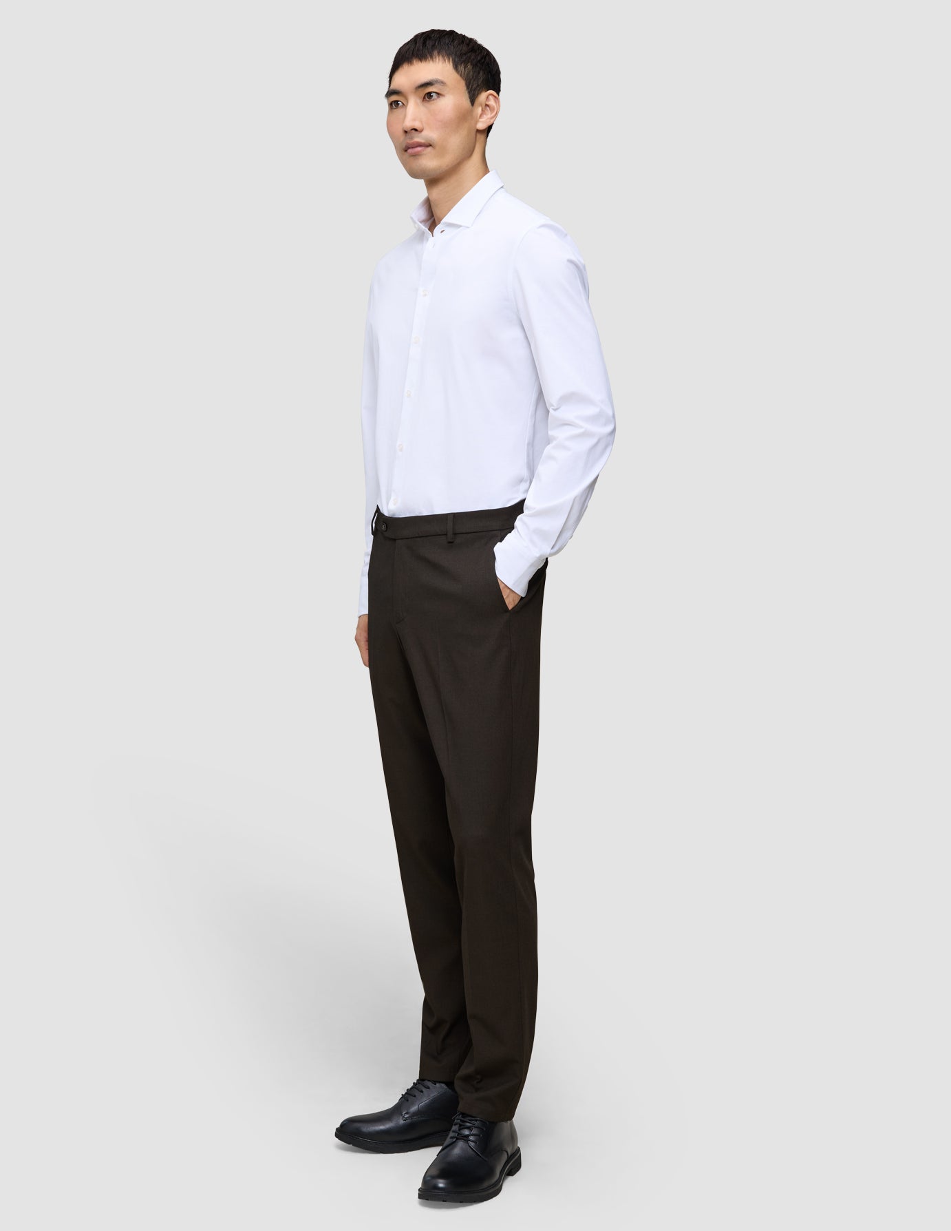 En mand i en hvid skjorte og et par Motion Suit-bukser i farven Regular Cognac med 360°-stretch, der står med hånden i lommen.